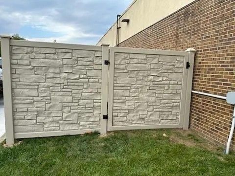 Gray faux-stone fence with a gate in front of a brick building and green grass.