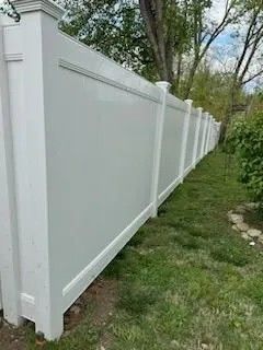 White vinyl privacy fence in a grassy yard, partially obscured by trees.