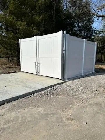 White fenced enclosure with gated doors on a concrete pad, gravel ground, trees in the background.