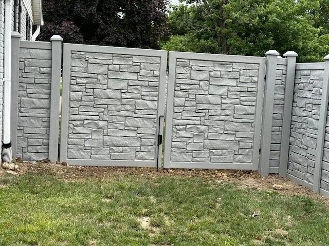 Gray stone-textured concrete gate and fence in a grassy yard, with a partly visible house and trees in the background.