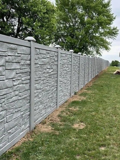 Gray stone-textured concrete fence in a grassy yard, with trees in the background under a cloudy sky.