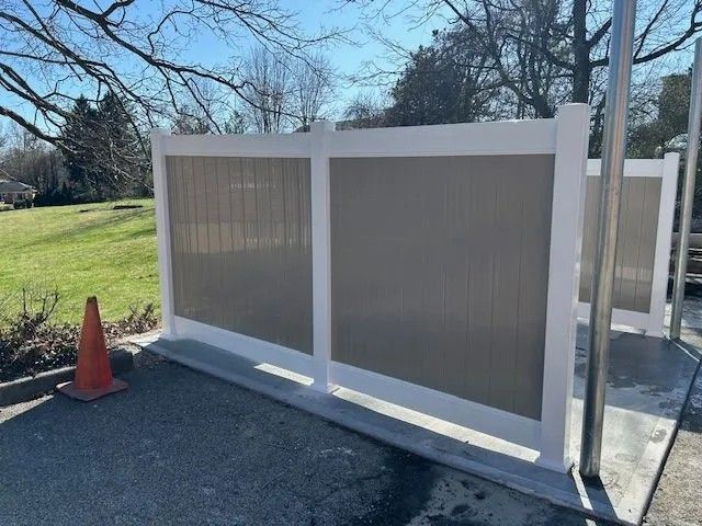 White and tan privacy fence panels on concrete base, with a safety cone in front, outdoors.