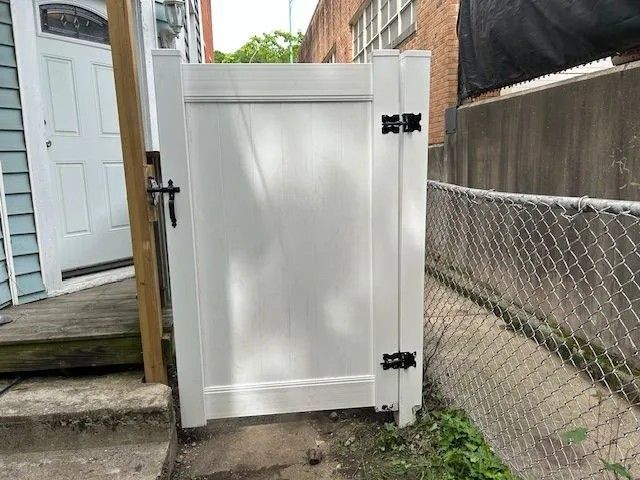 White vinyl gate with black hardware, next to a chain-link fence and concrete wall, leading to a door.