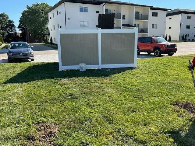 A tan and white fence stands on a grassy lawn with a building and cars in the background.