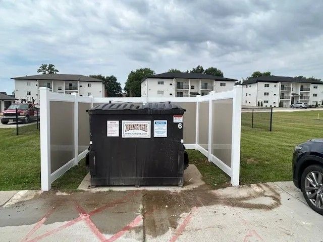 Black dumpster enclosed by a white fence on a grassy area, in front of apartment buildings under a cloudy sky.