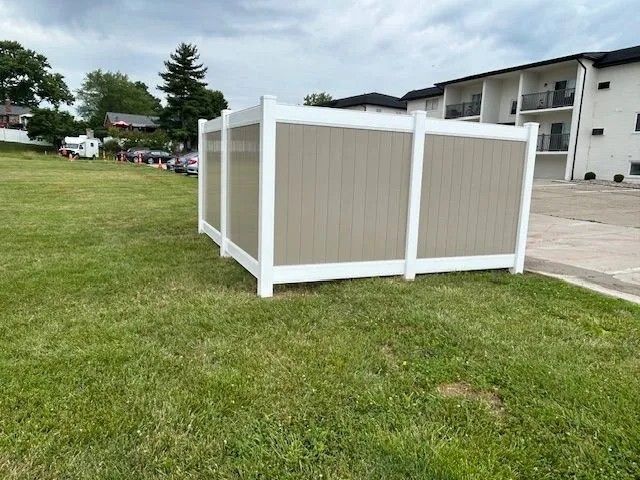 A tan and white vinyl fence on a grassy lawn, with a multi-story building in the background.