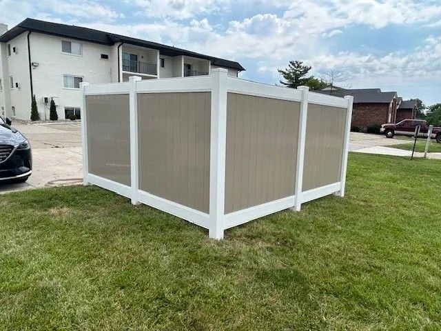 A tan and white vinyl fence encloses a small area on a grassy lawn, with a building and cars in the background.