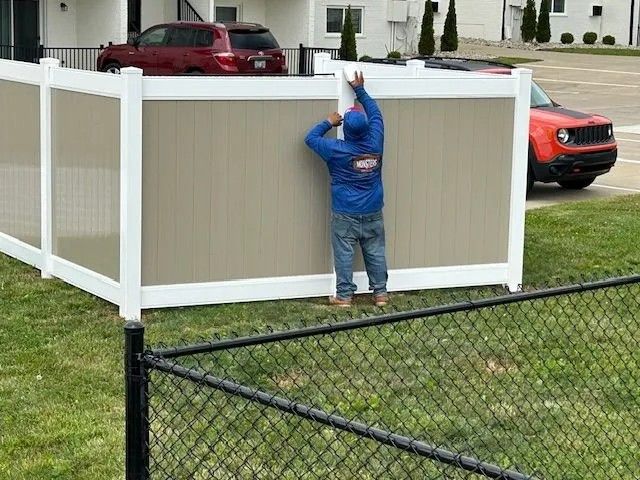 Person installing a tan and white privacy fence outdoors. A red SUV and orange Jeep are parked nearby.