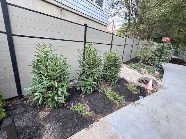 Beige and black fence with green shrubbery and a concrete sidewalk.