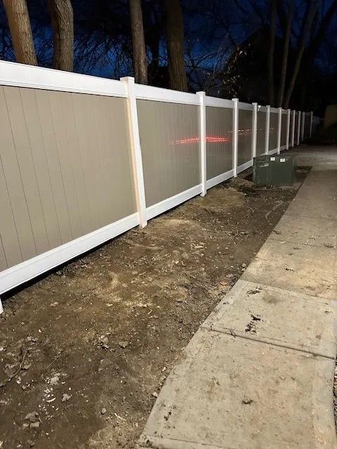 Tan and white vinyl fence along a dirt area beside a sidewalk, trees in background, dusk.