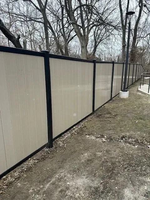 Beige vertical panel fence with black posts, extending along a grassy area, trees in the background.