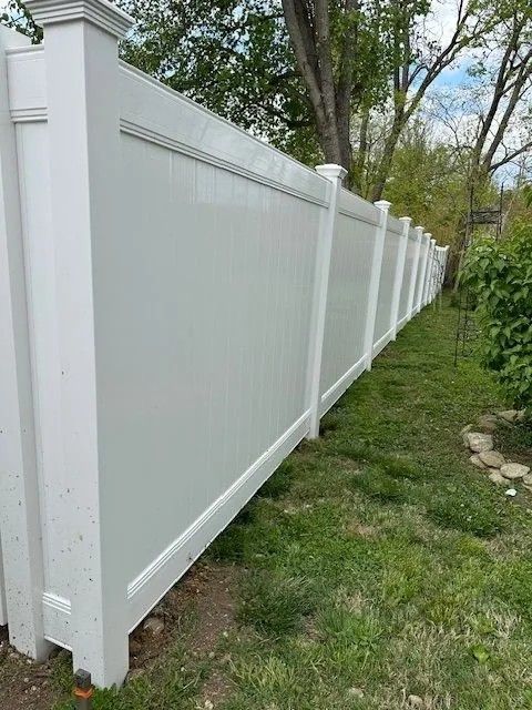 White vinyl privacy fence in a grassy yard, partially obscured by trees.