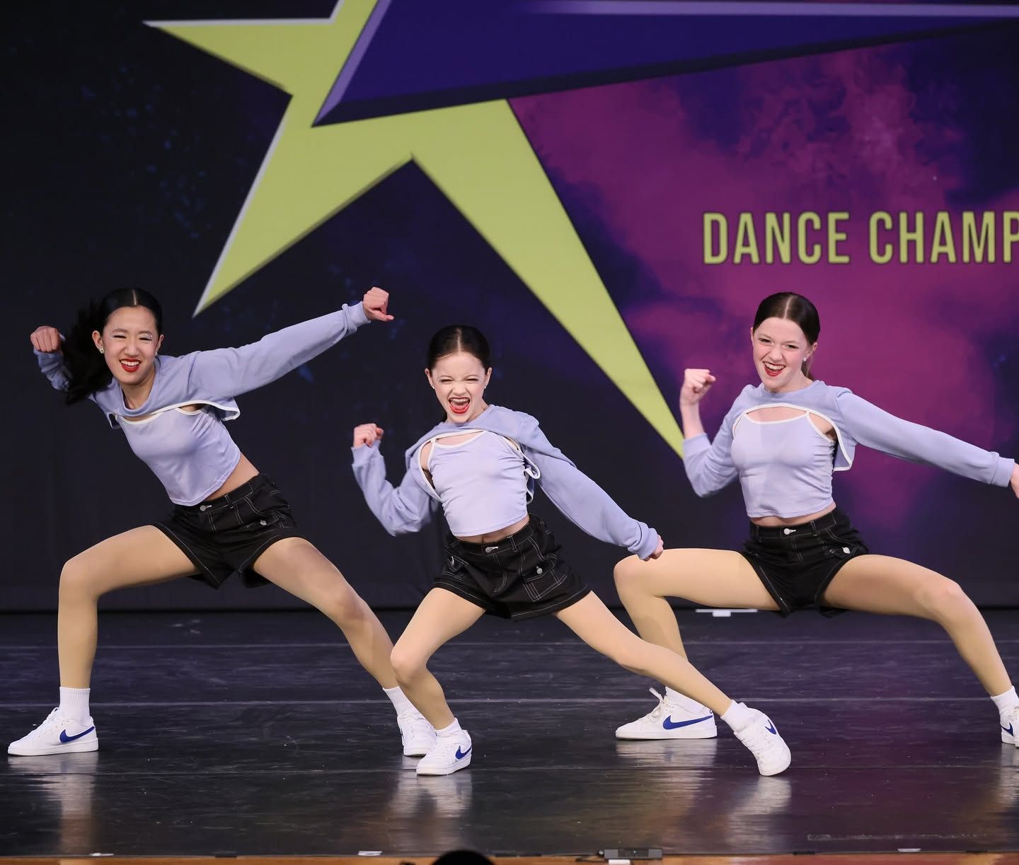 Three dancers in gray tops and black shorts pose onstage before a purple dance competition backdrop.