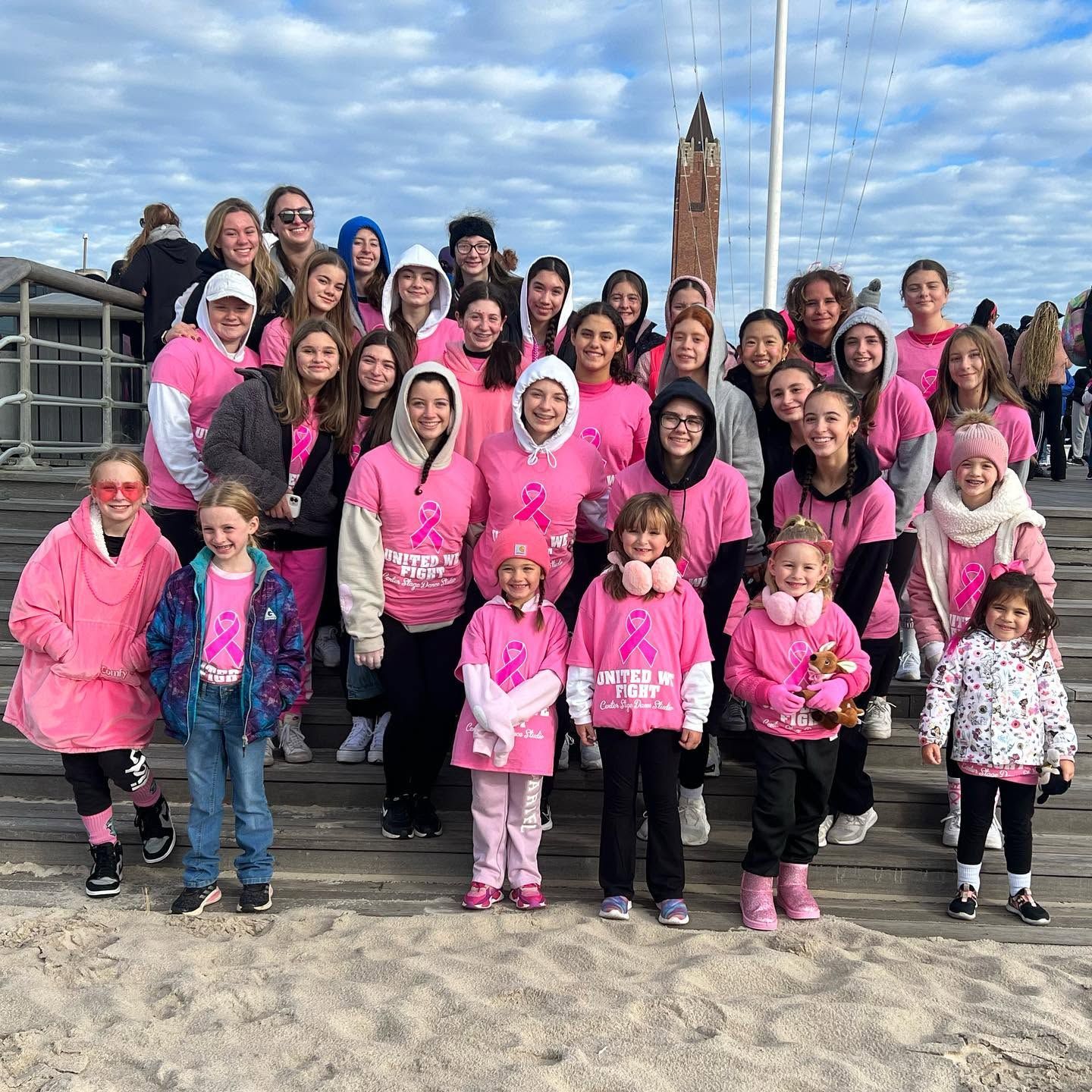 Large group in pink shirts posing on steps outdoors with a tall stone monument behind them