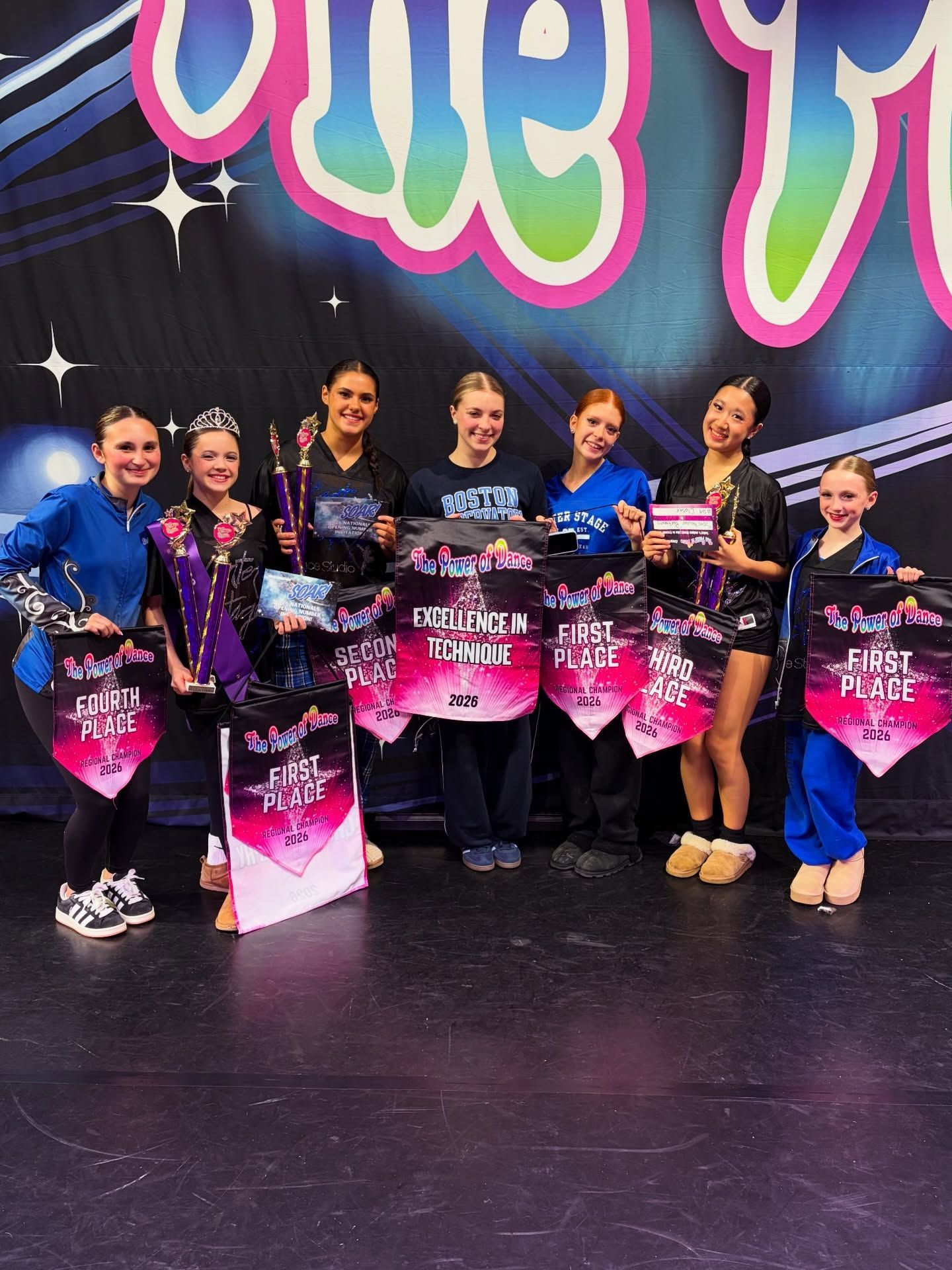 Group of dancers posing onstage with pink trophy bags in front of a colorful backdrop