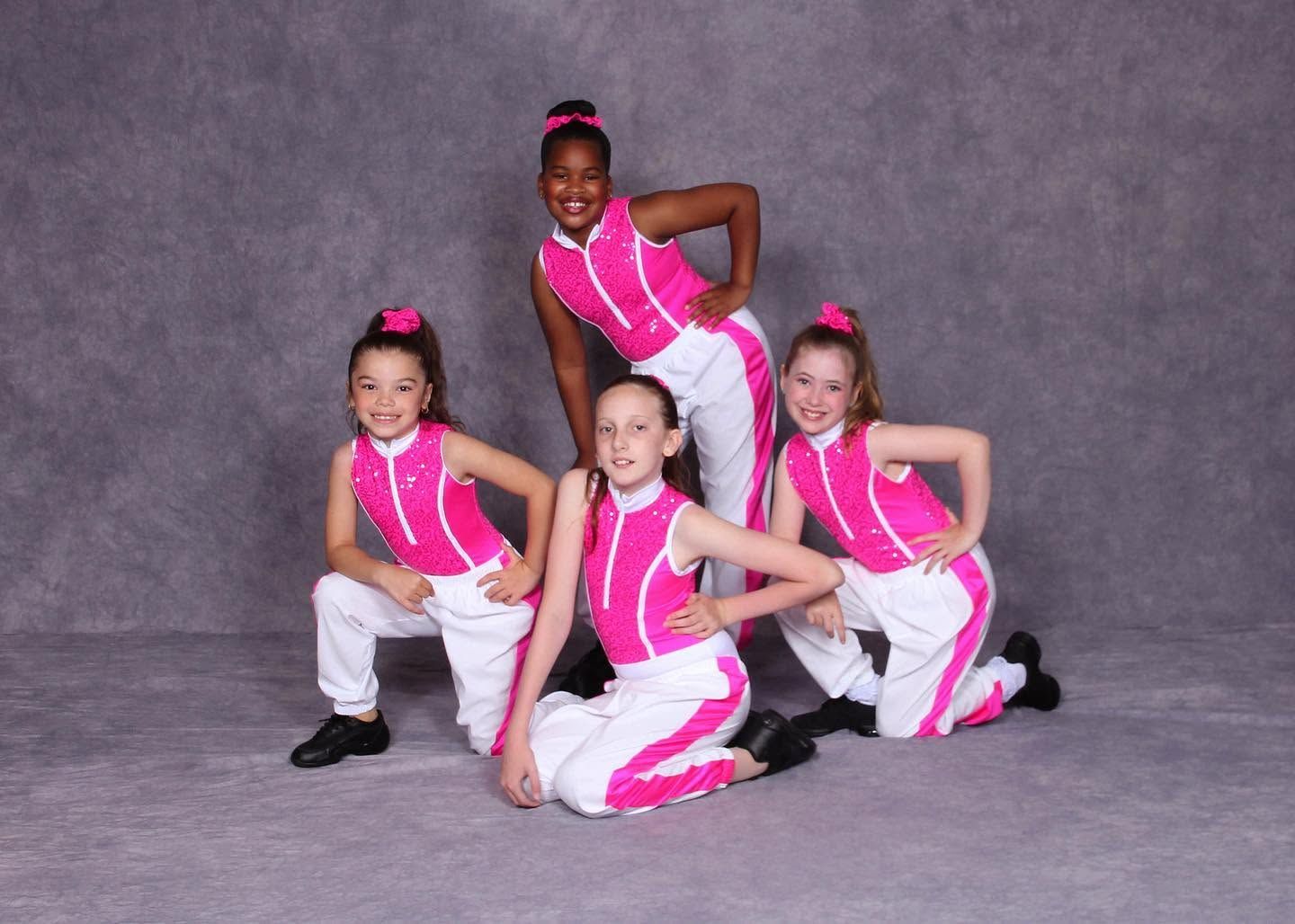 Four children in pink dance costumes posing in a studio against a gray backdrop
