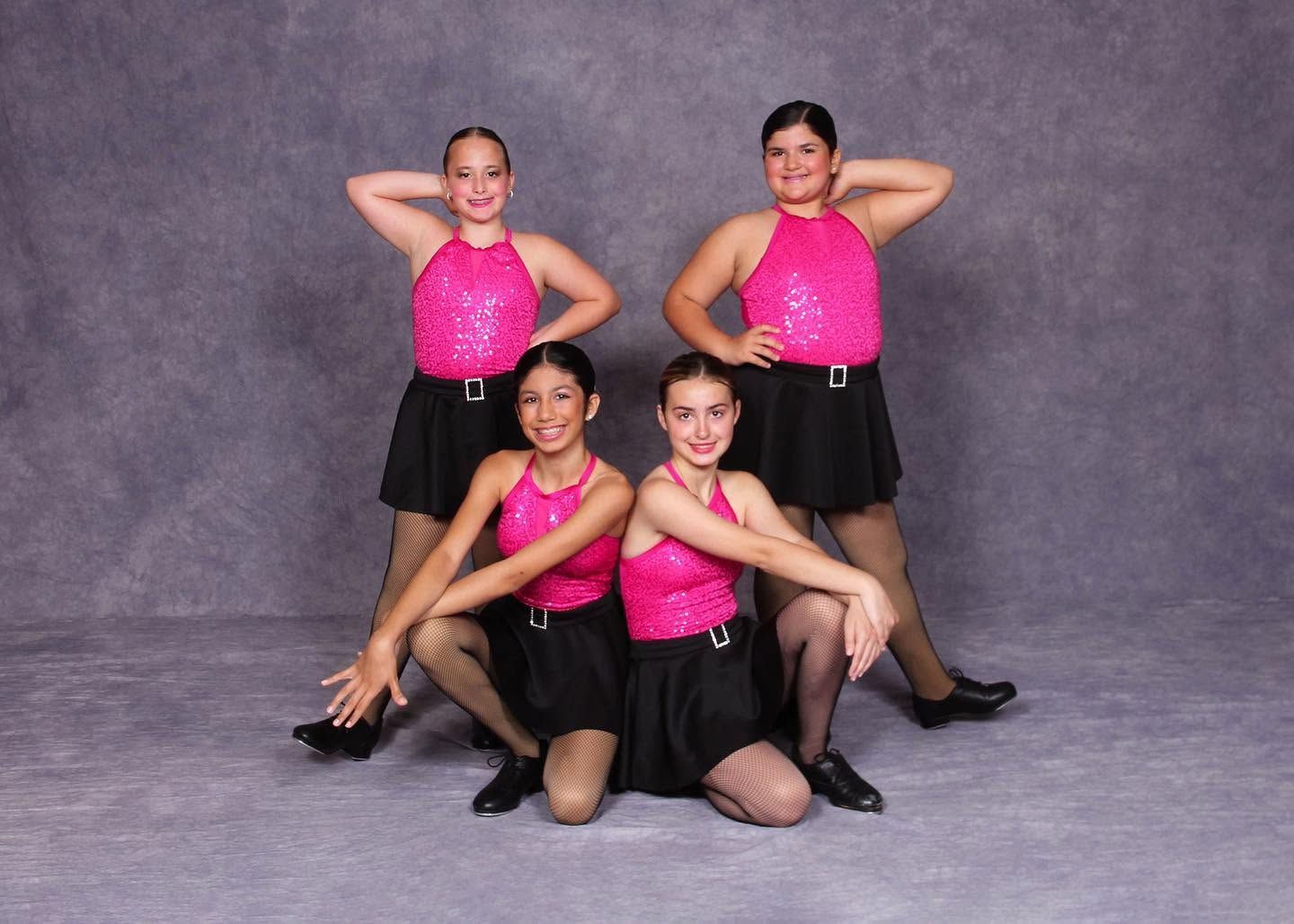 Four dancers in bright pink costumes pose in a studio, two standing behind and two kneeling in front.