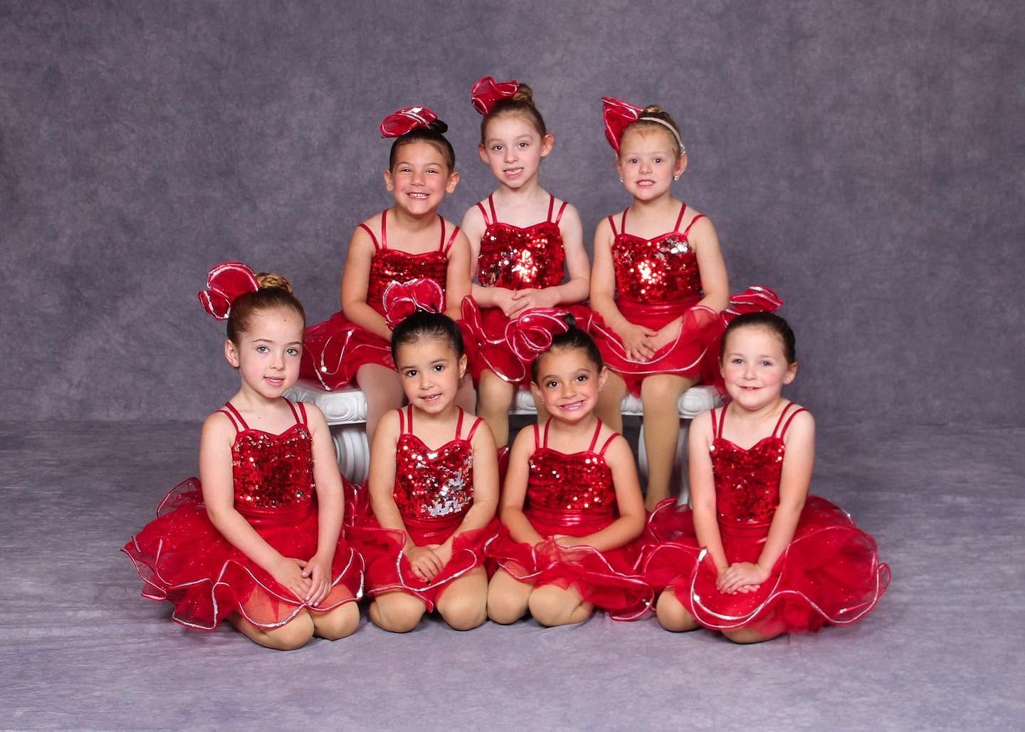 Seven children in red dance costumes posing against a gray studio backdrop.