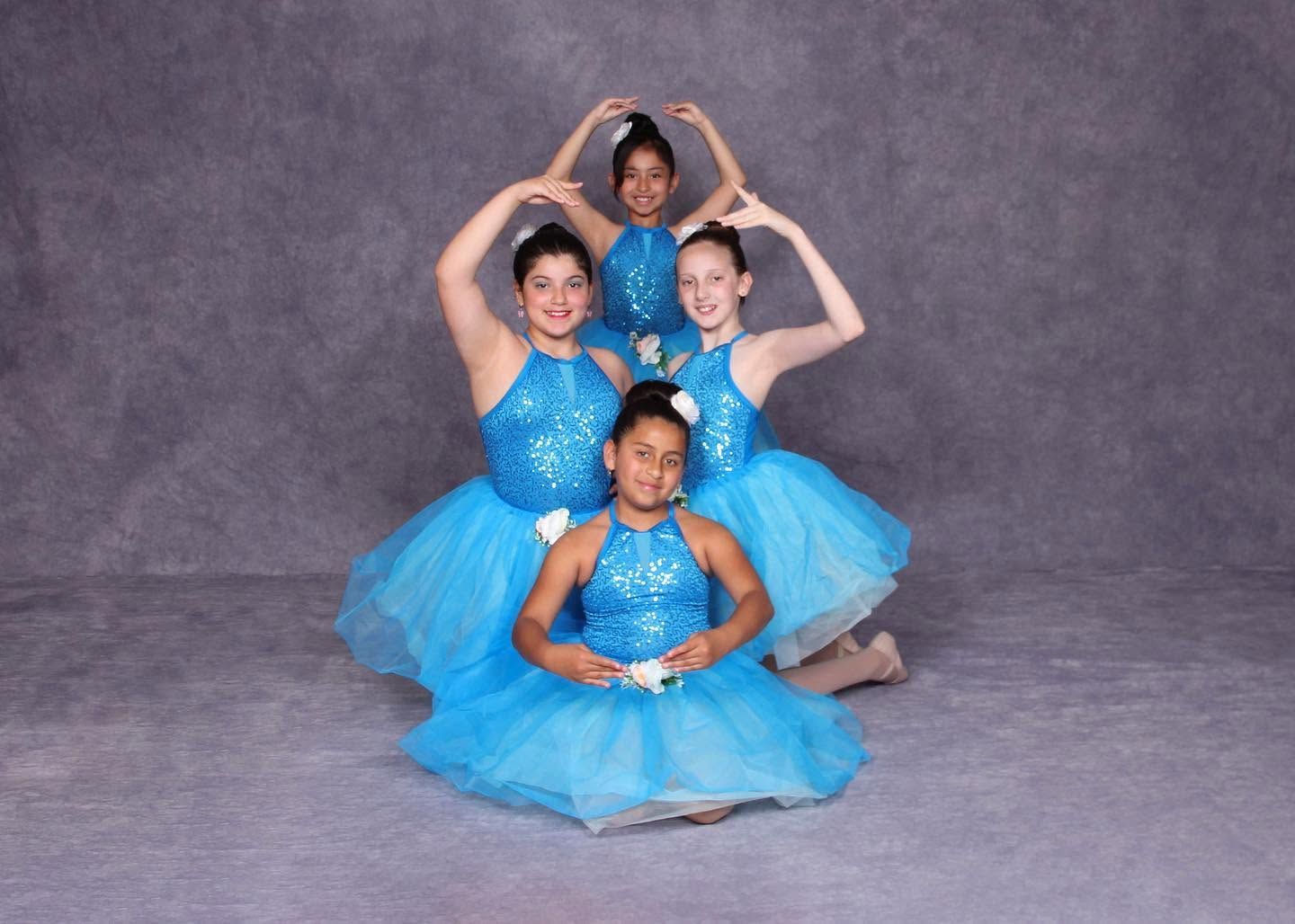 Four dancers in blue tutus pose in a stacked formation against a gray studio backdrop.