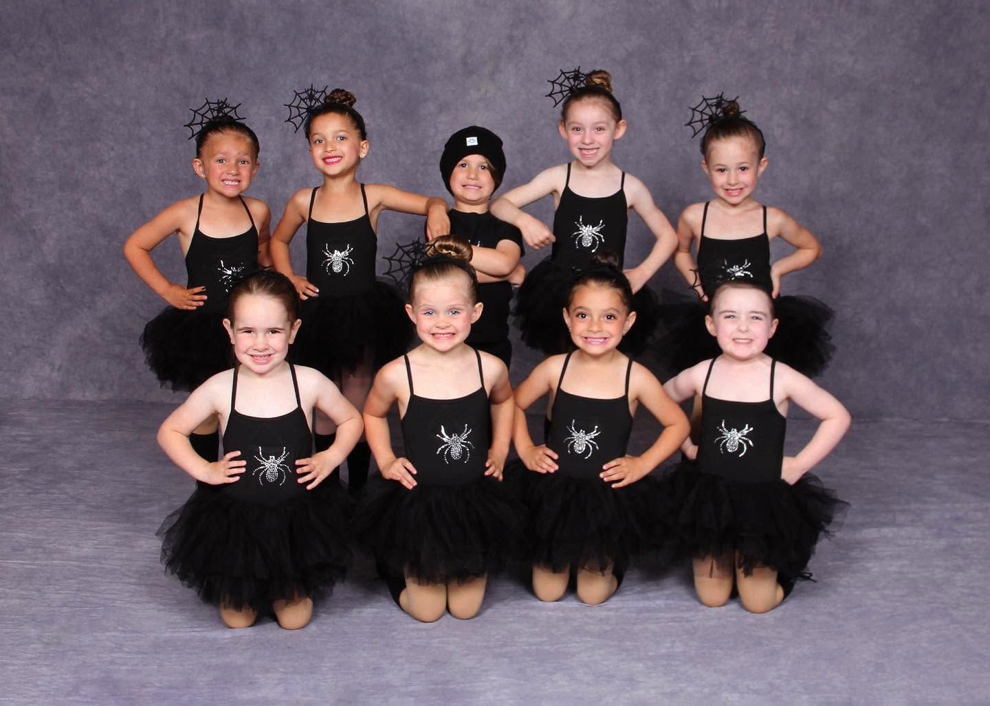 Nine young ballet dancers in black tutus pose in two rows against a gray studio backdrop.