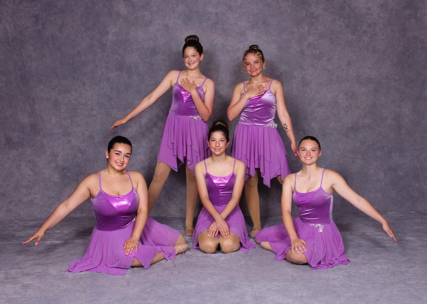 Five dancers in purple costumes pose on a gray studio backdrop, three standing and two kneeling.