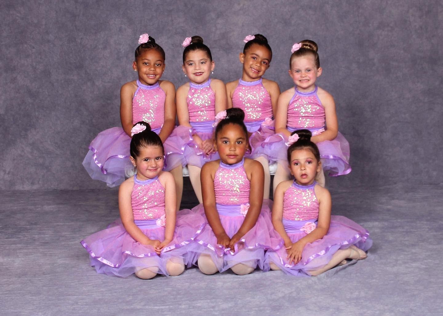 Seven children in matching purple ballet costumes posed in two rows against a gray backdrop.