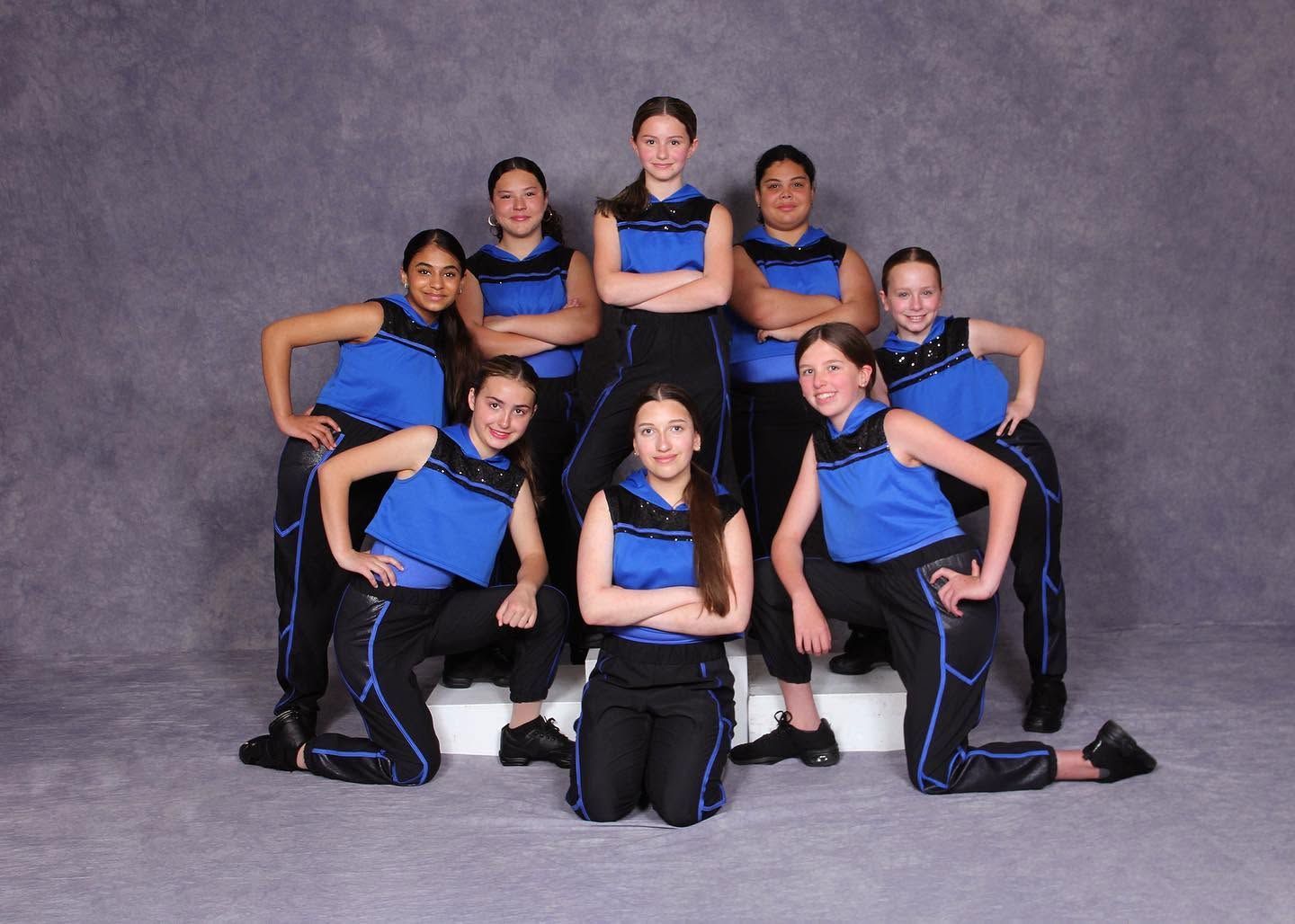 Eight dancers in blue and black costumes pose in a studio, arranged in a pyramid formation.