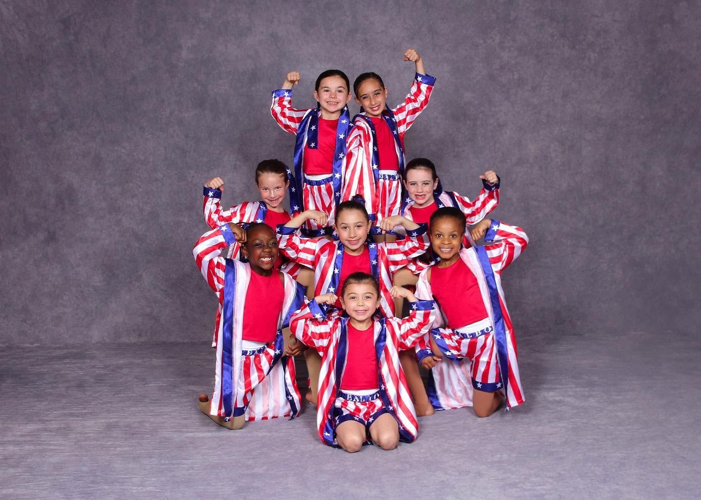 Eight children in red, white, and blue costumes pose in a pyramid formation against a gray backdrop.