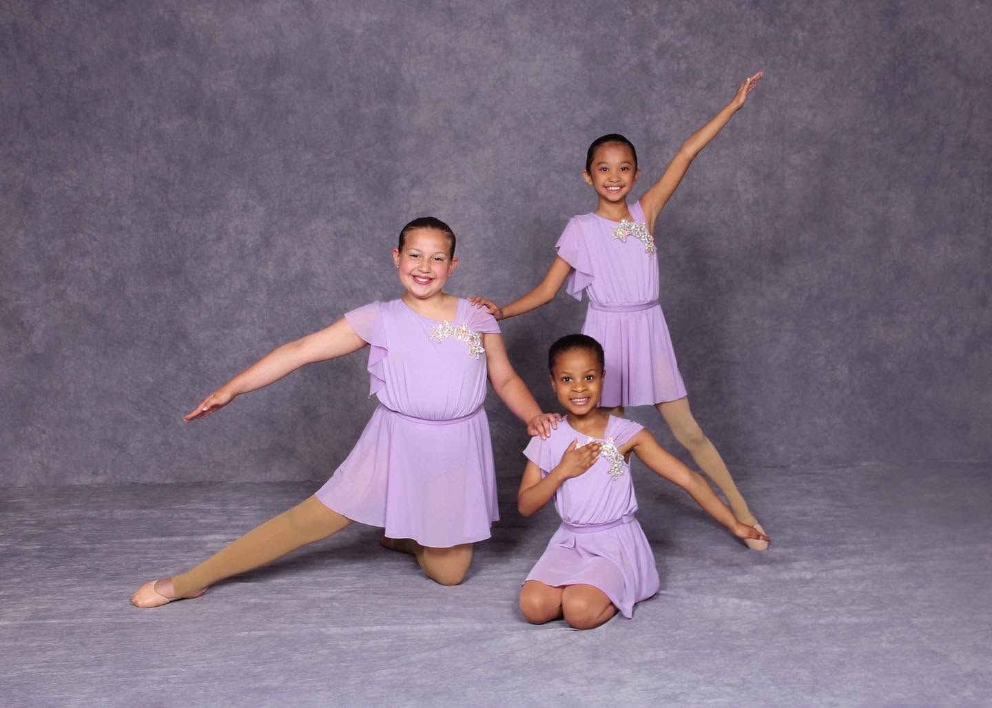 Three dancers in lavender costumes pose on a gray studio backdrop.