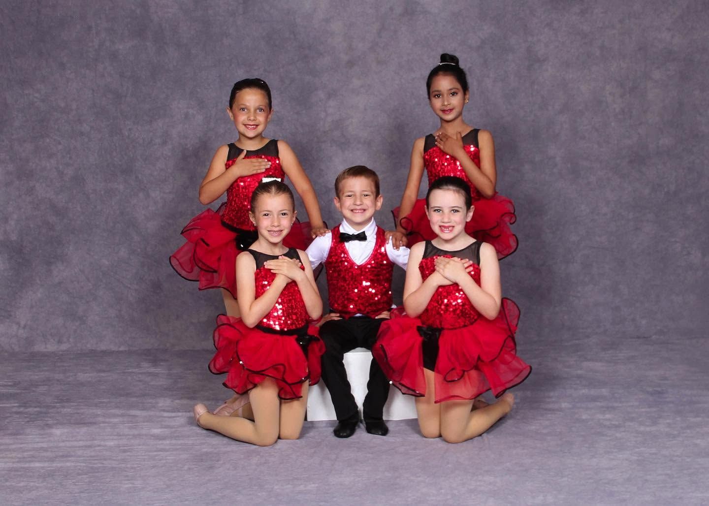 Five young dancers in red costumes posing in a studio, with one seated center and four kneeling around them