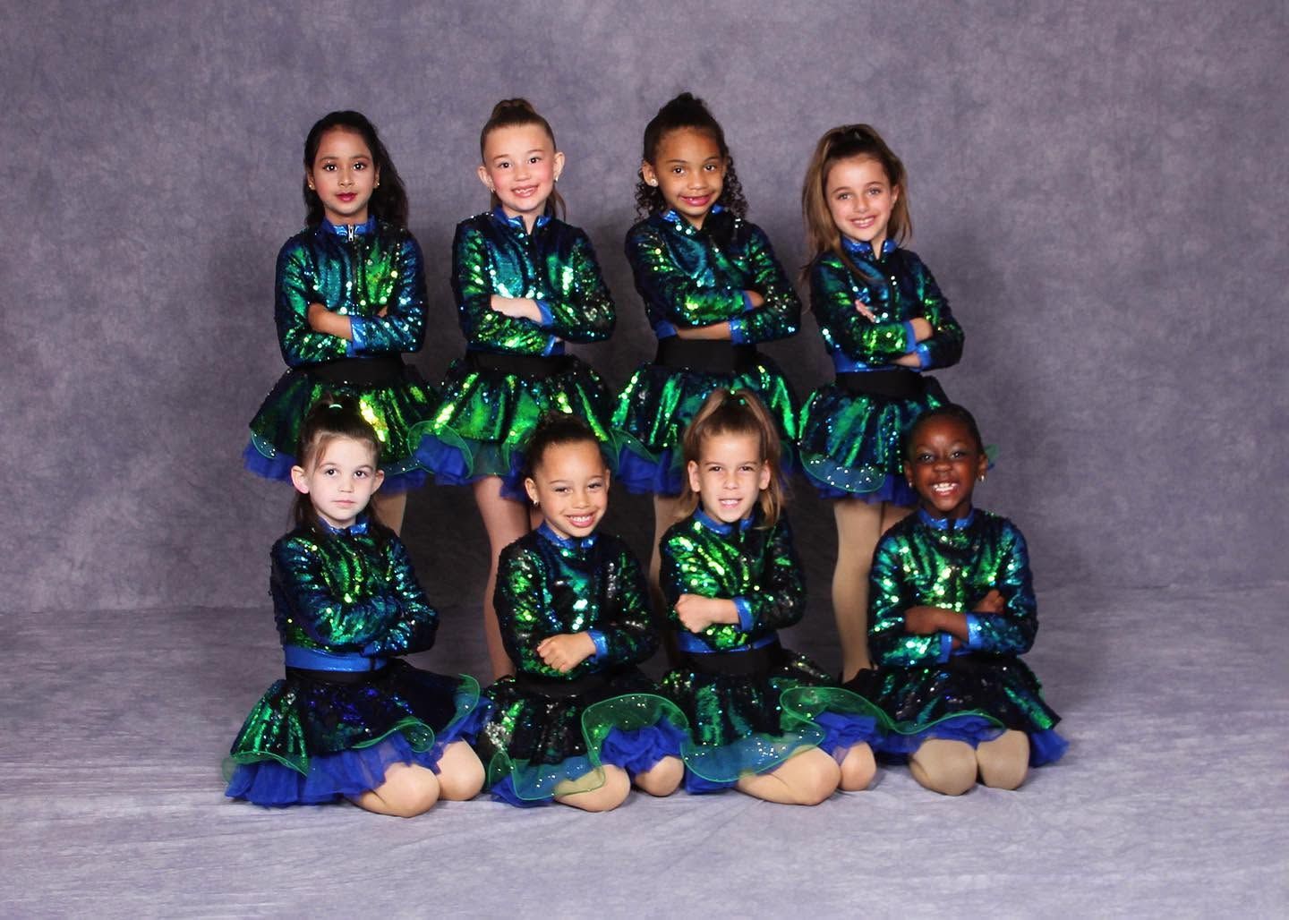 Seven children in shimmering green-and-blue dance costumes seated and kneeling against a gray studio backdrop