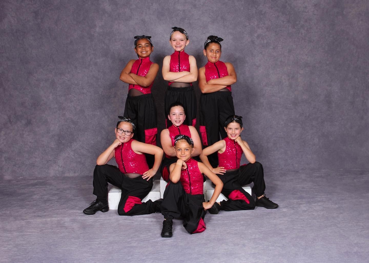 Seven dancers in red tops and black pants pose in a studio against a gray backdrop.