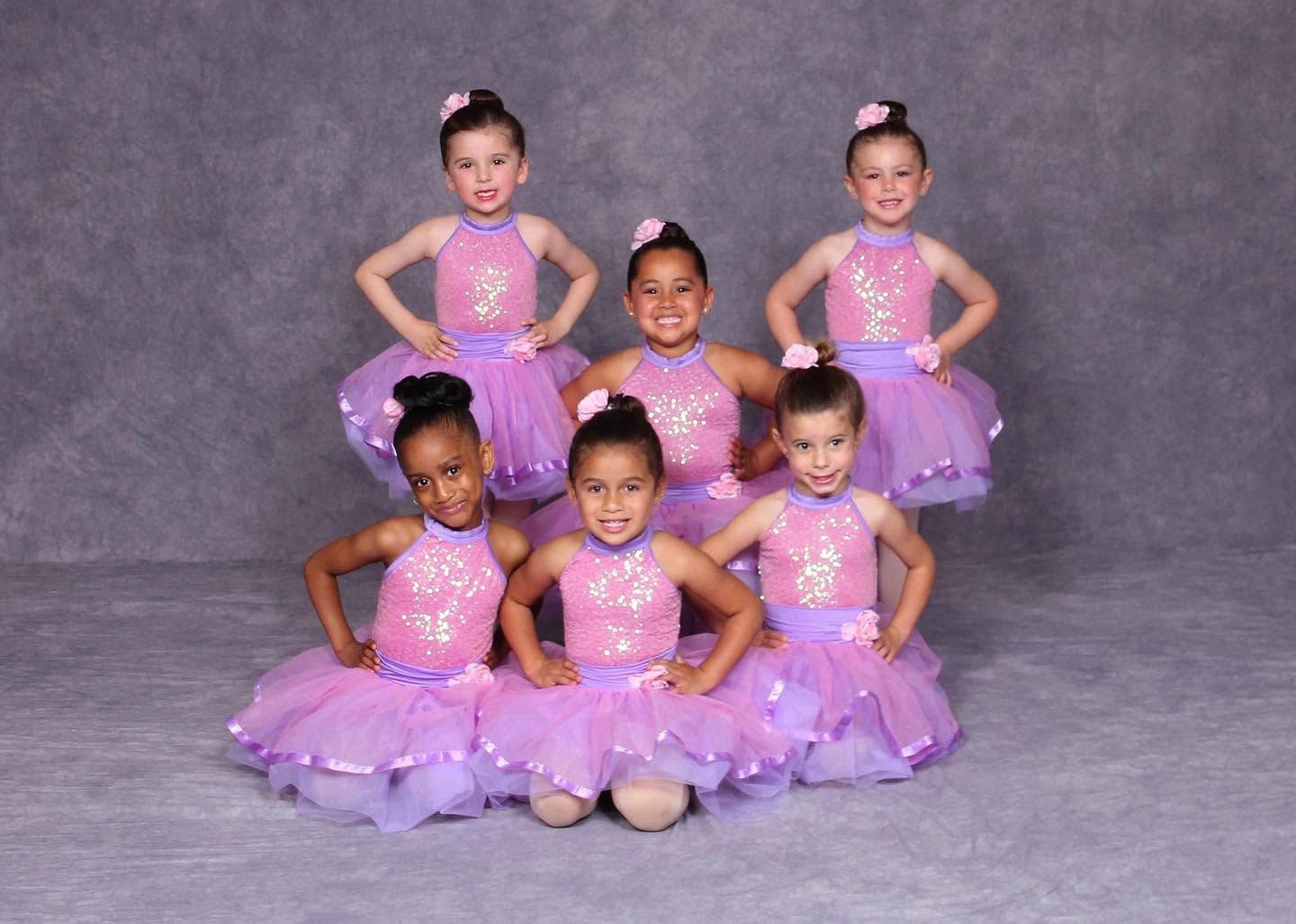 Six young dancers in pink-purple tutus posing against a gray studio backdrop
