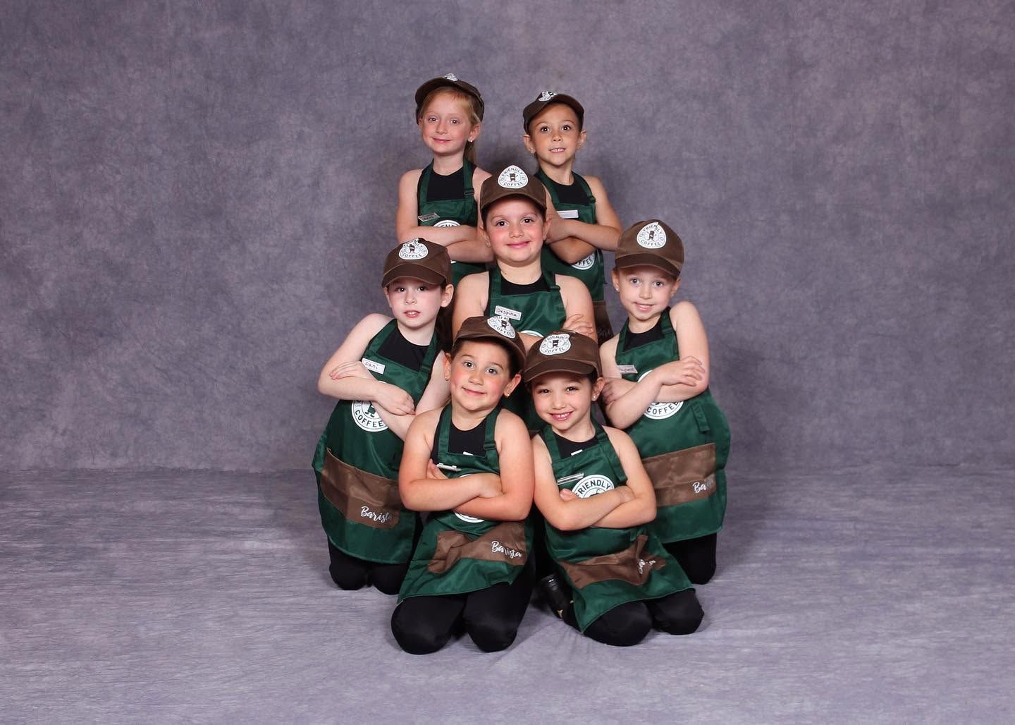 Group of young performers in green costumes posing in a stacked formation against a gray backdrop