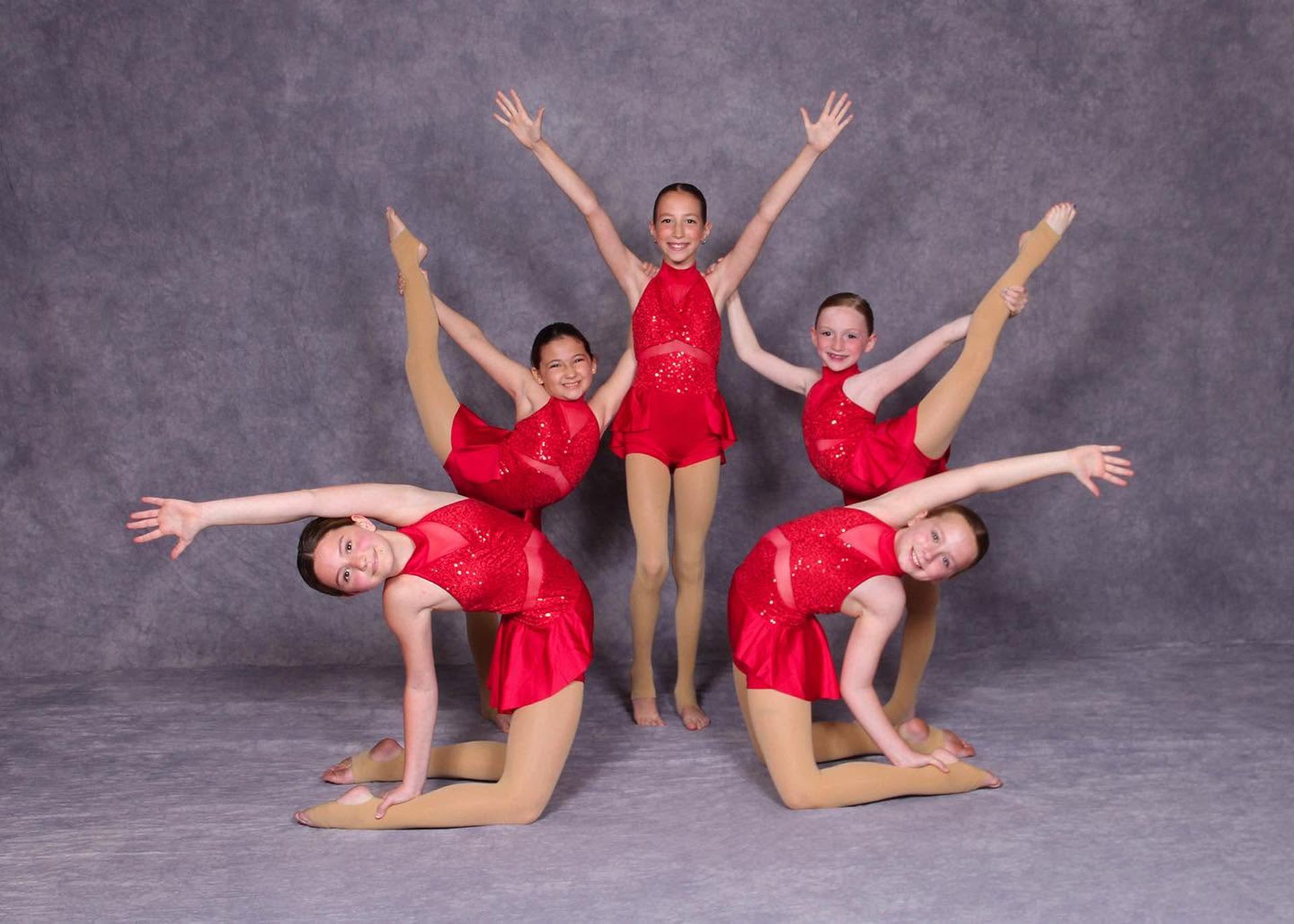 Five dancers in red leotards pose in a synchronized acrobatic formation on a gray backdrop.