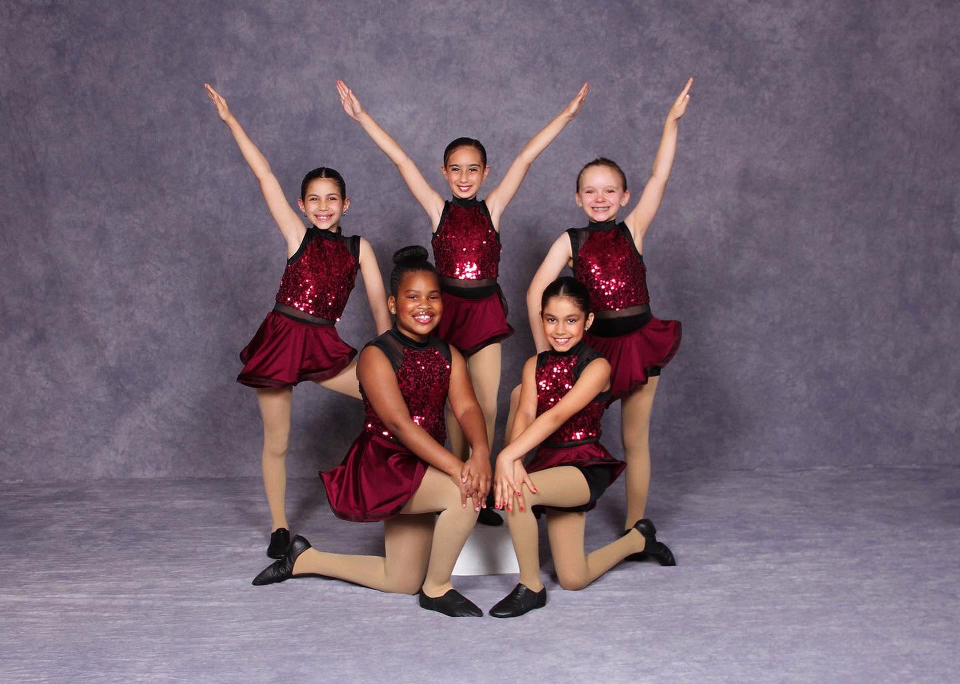 Five dancers in red sequined costumes pose on a studio backdrop, arms raised in a cheer formation.