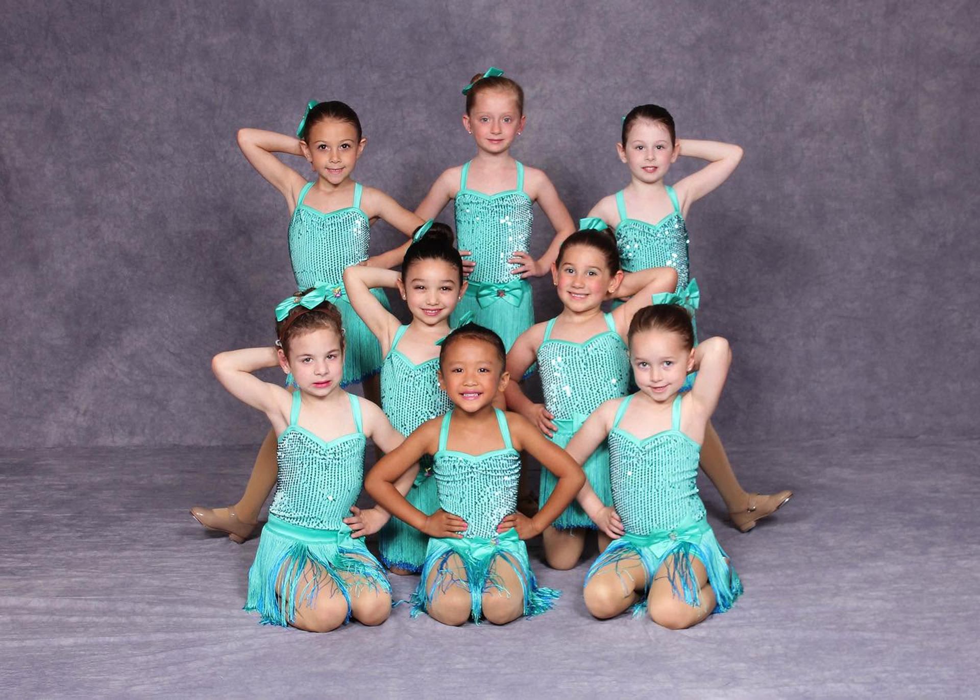 Seven young dancers in turquoise costumes pose in a studio with gray backdrop.