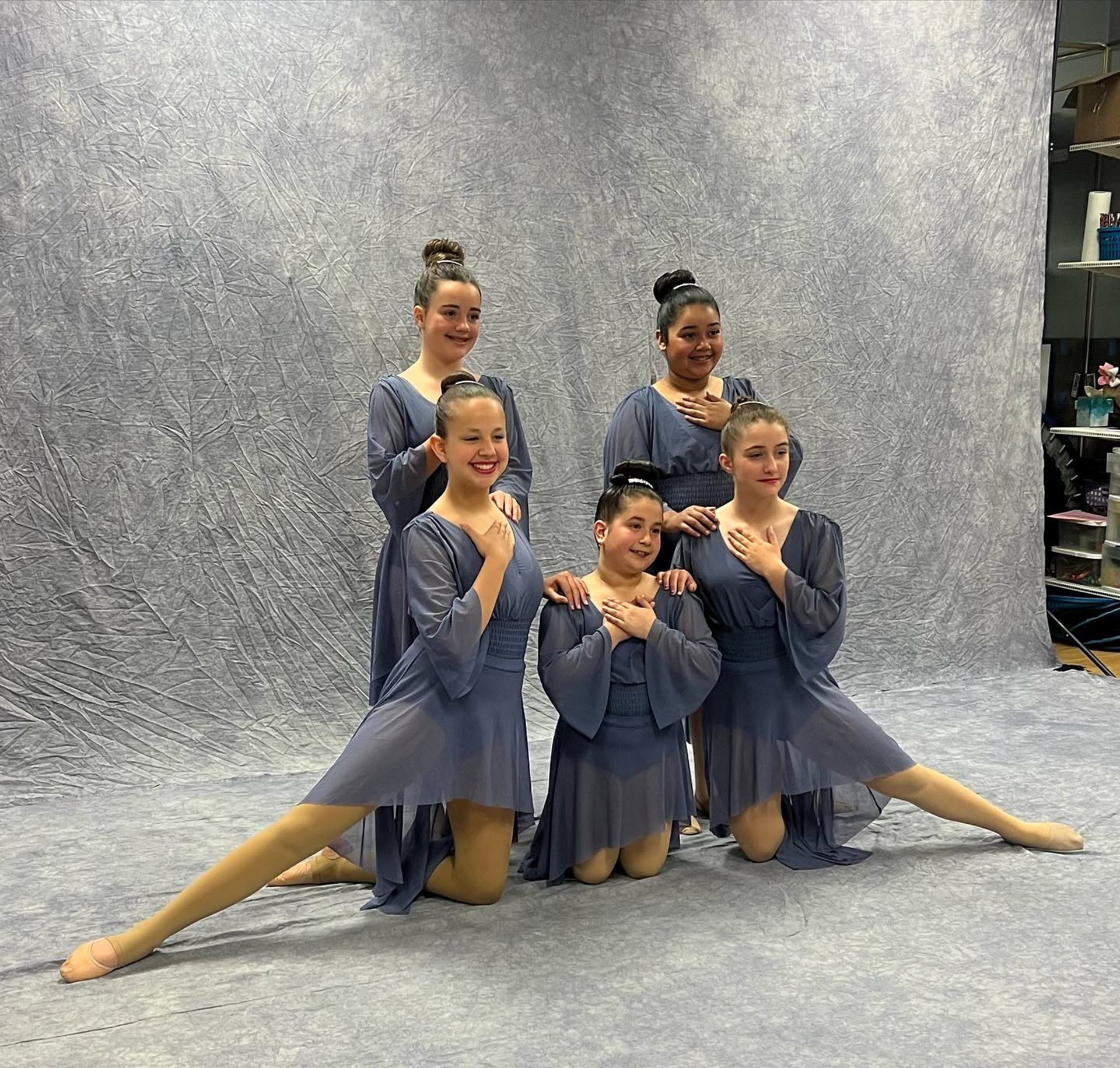Five dancers in blue costumes pose in a studio with a gray backdrop.