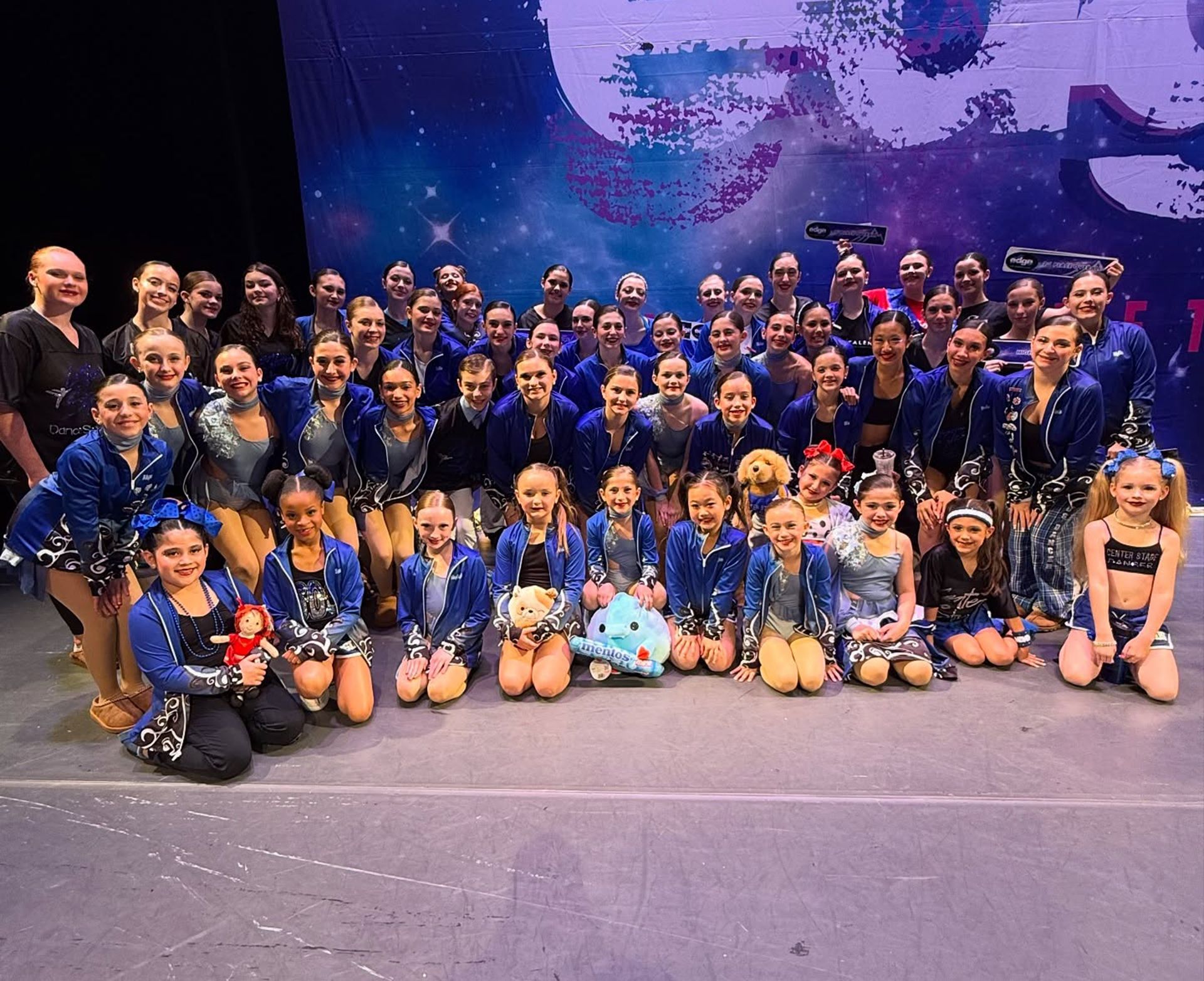 Large group posing onstage in matching blue costumes against a starry purple backdrop.