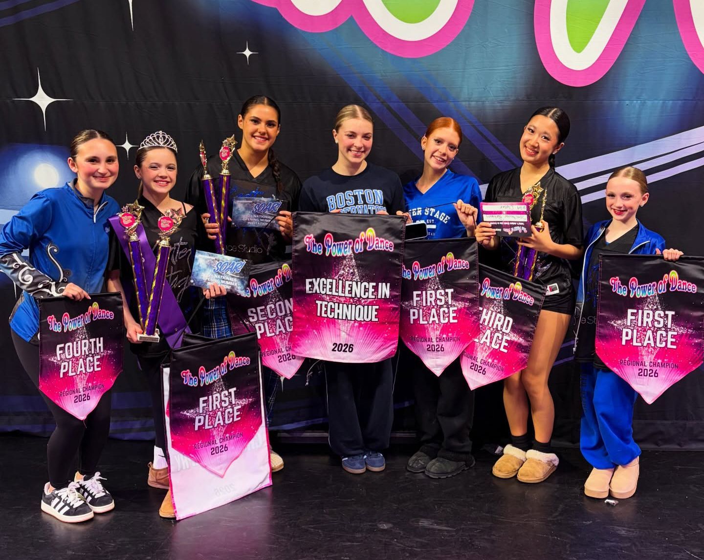 Seven dancers posing on a stage with pink first-place trophies and black outfits, smiling at the camera.