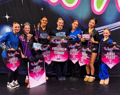 Seven dancers posing on a stage with pink first-place trophies and black outfits, smiling at the camera.