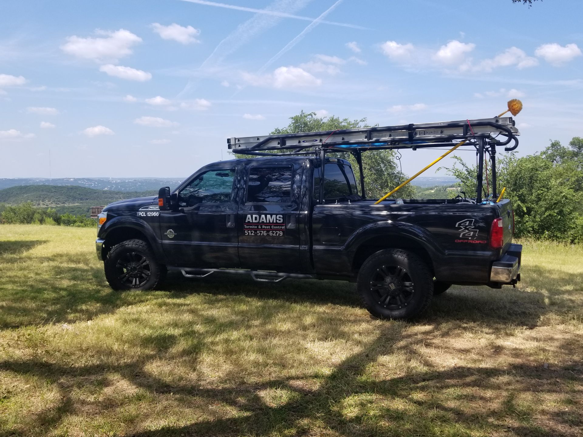 Black work truck with ladder rack parked on a grassy hillside under a blue sky.