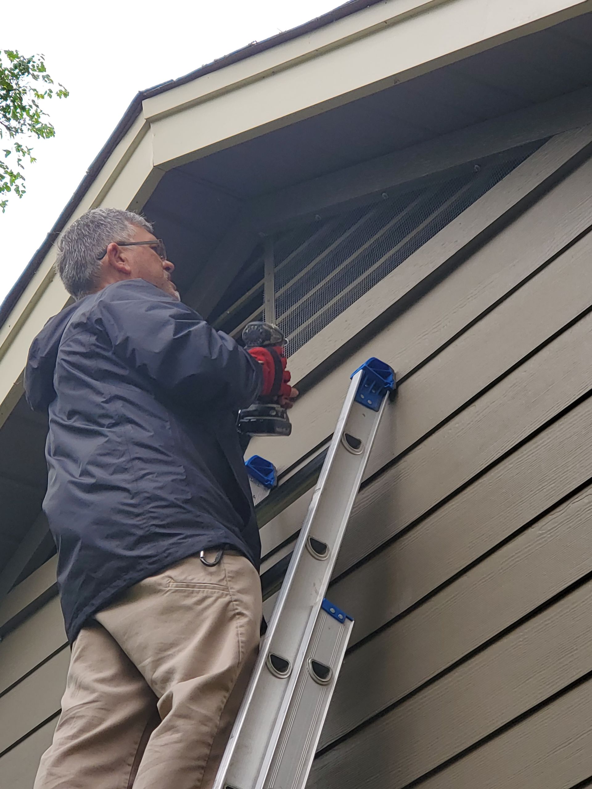 Man on ladder using drill on siding near a triangular vent. Building exterior.