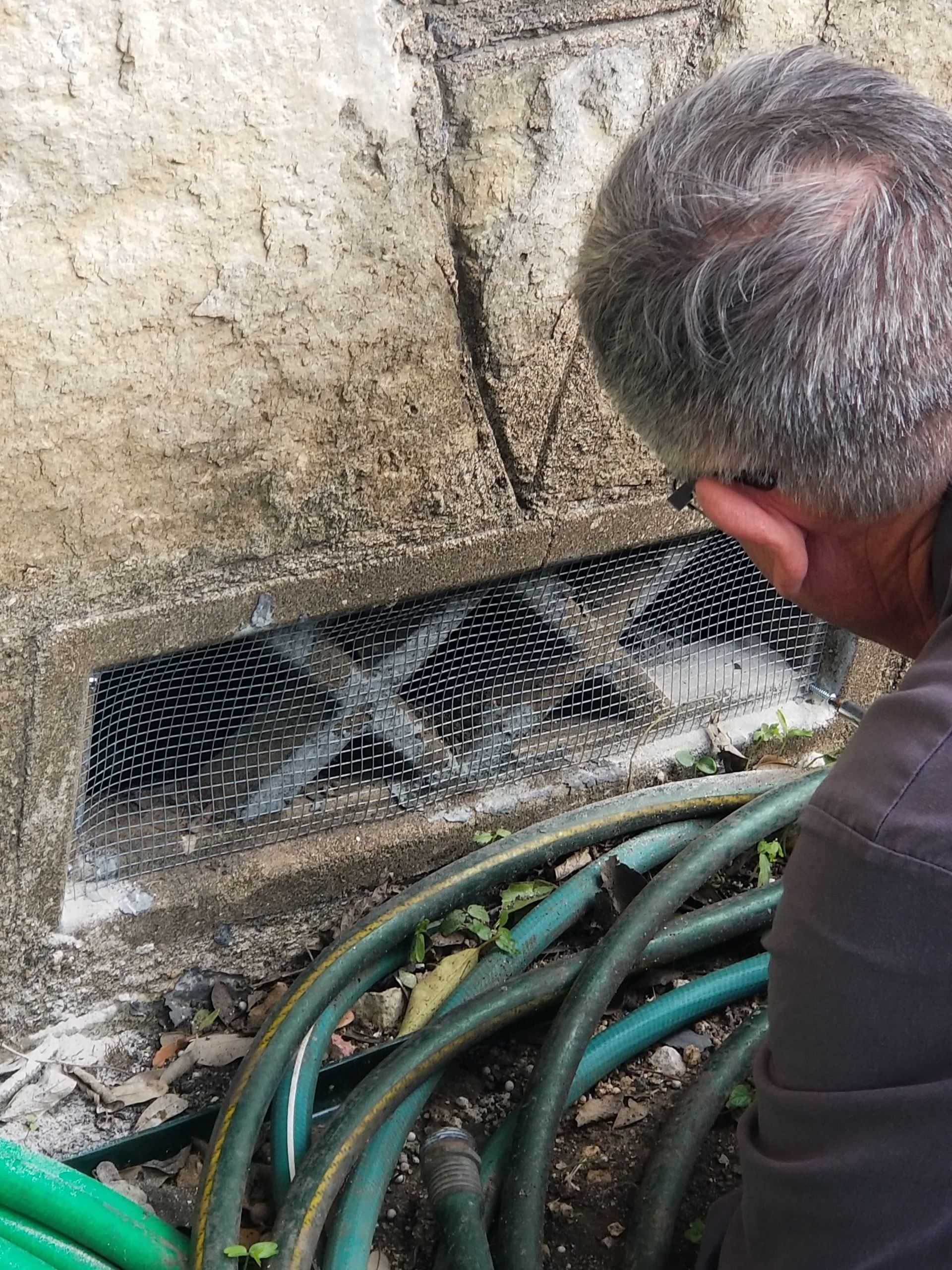 Man looking at a vent covered with a mesh on an exterior wall. Green hose on ground below.