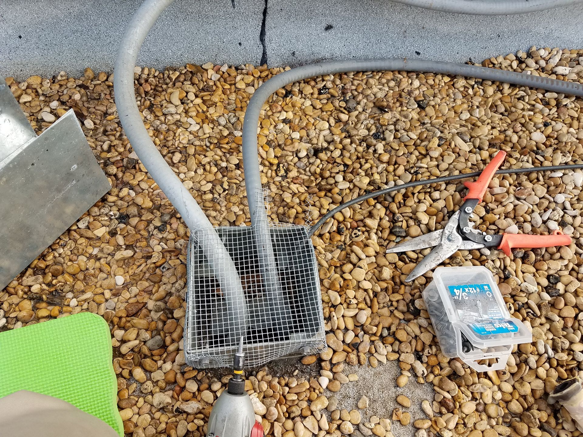 A metal drainage grate with three gray tubes connected, surrounded by gravel, tools, and a green pad.
