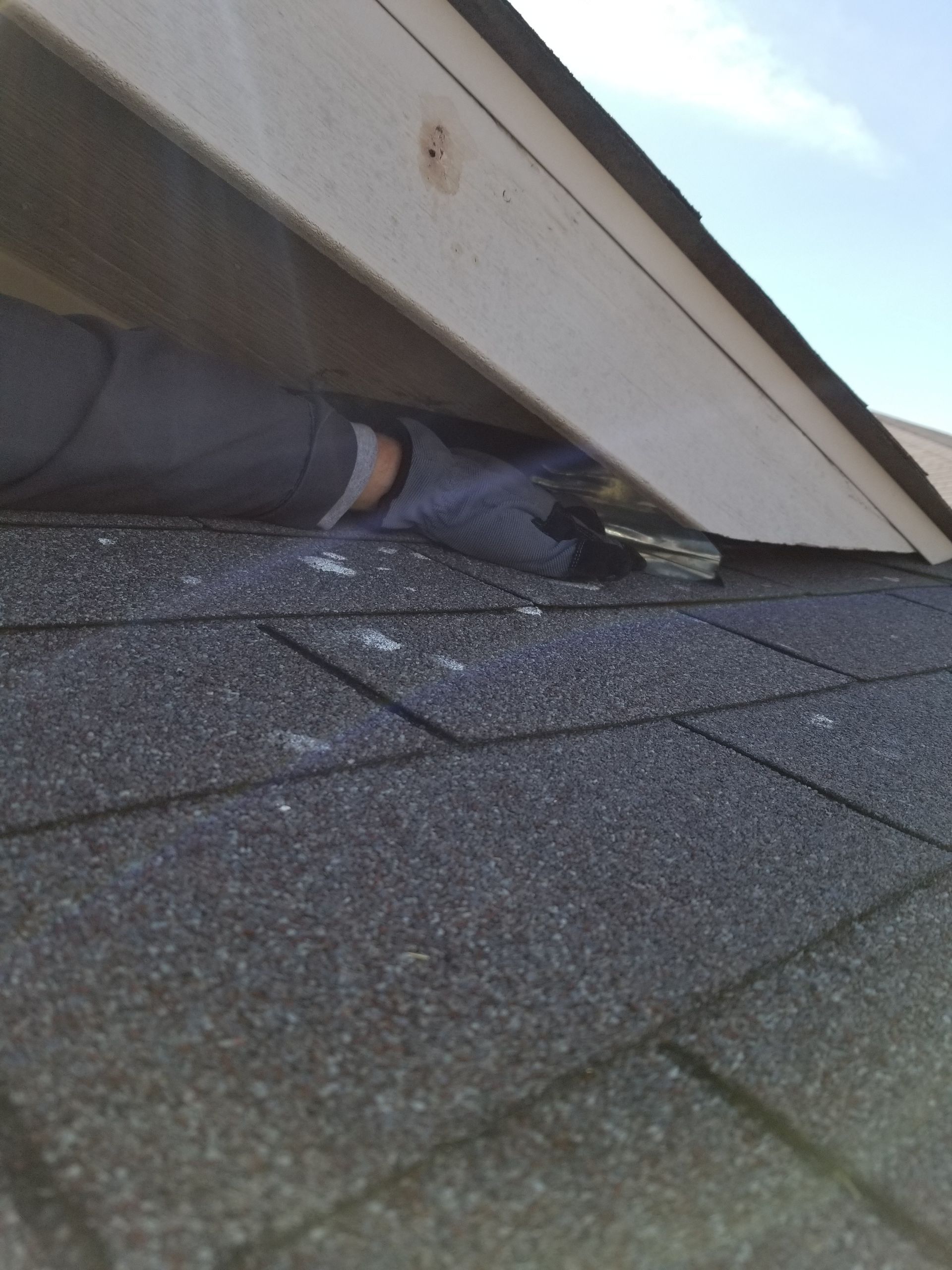 Person's gloved hand reaches under a roof overhang, near asphalt shingles.