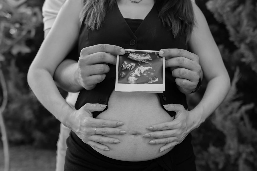A pregnant person holding an ultrasound photo over their belly with their hands on their abdomen outdoors