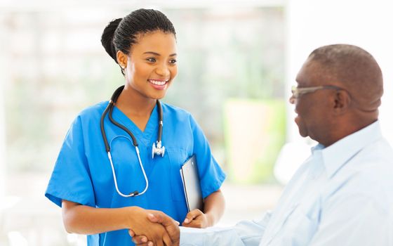 A smiling nurse in blue scrubs talks with a seated patient in a bright clinic room.
