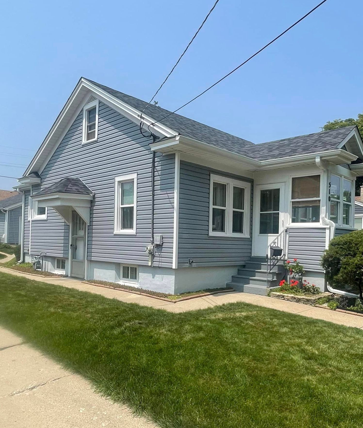 A small gray house with a gray roof is sitting on a lush green lawn next to a sidewalk