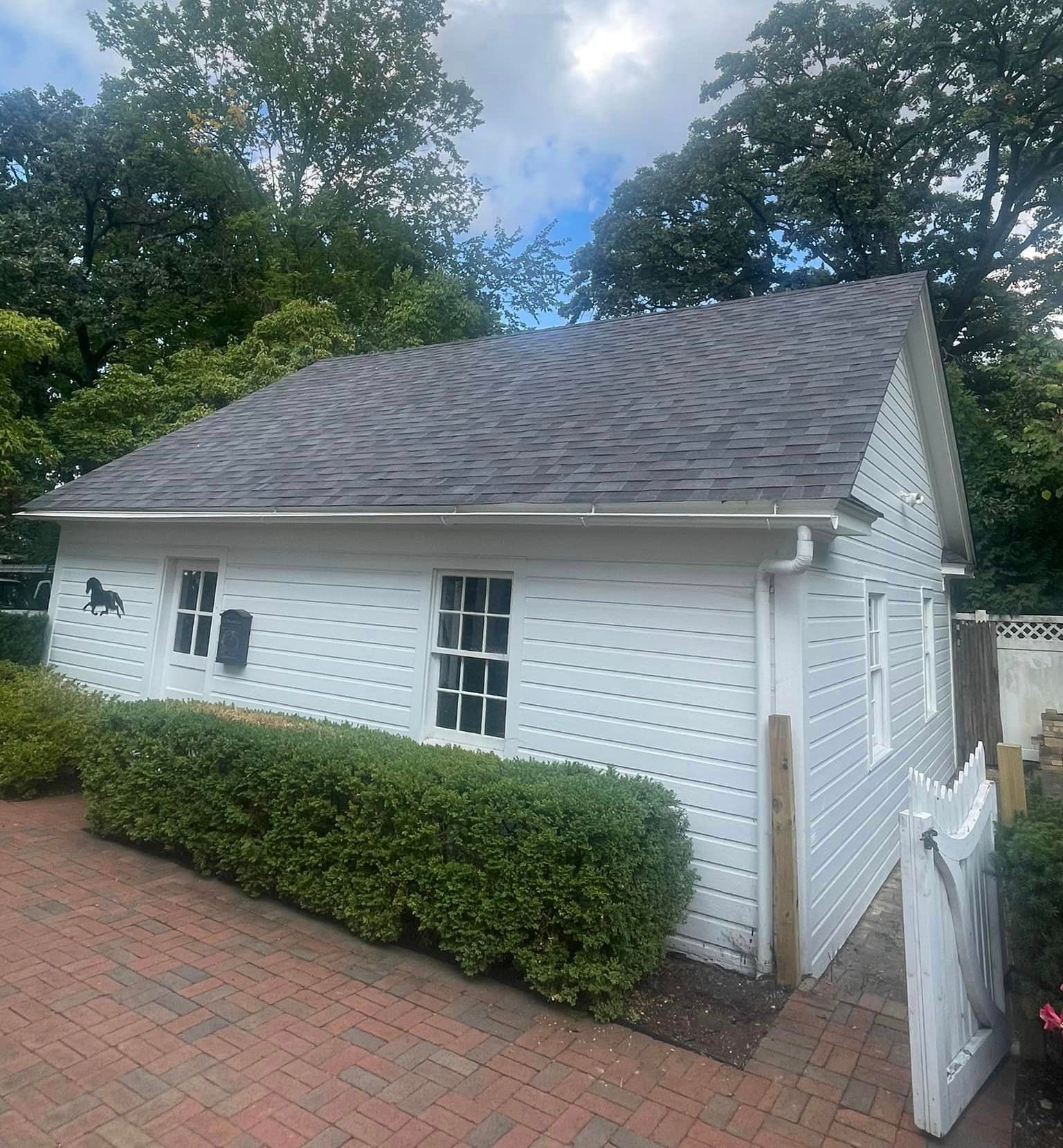A white house with a gray roof is surrounded by trees and bushes